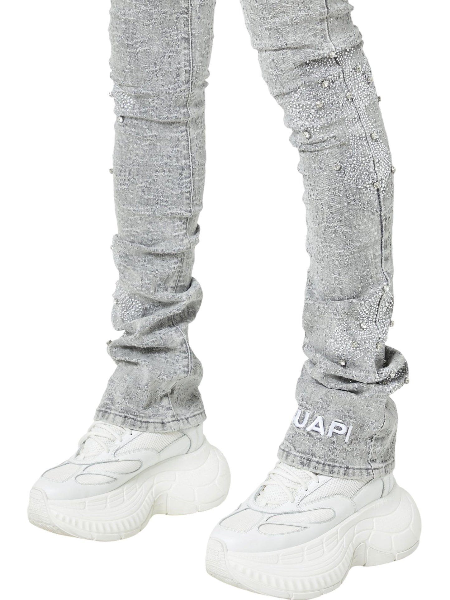 Person wearing GUAPI Smoke Grey Distressed Cross Denim and white chunky sneakers, standing with feet slightly apart. The jeans feature textured details and "GUAPI" branding near the ankle.