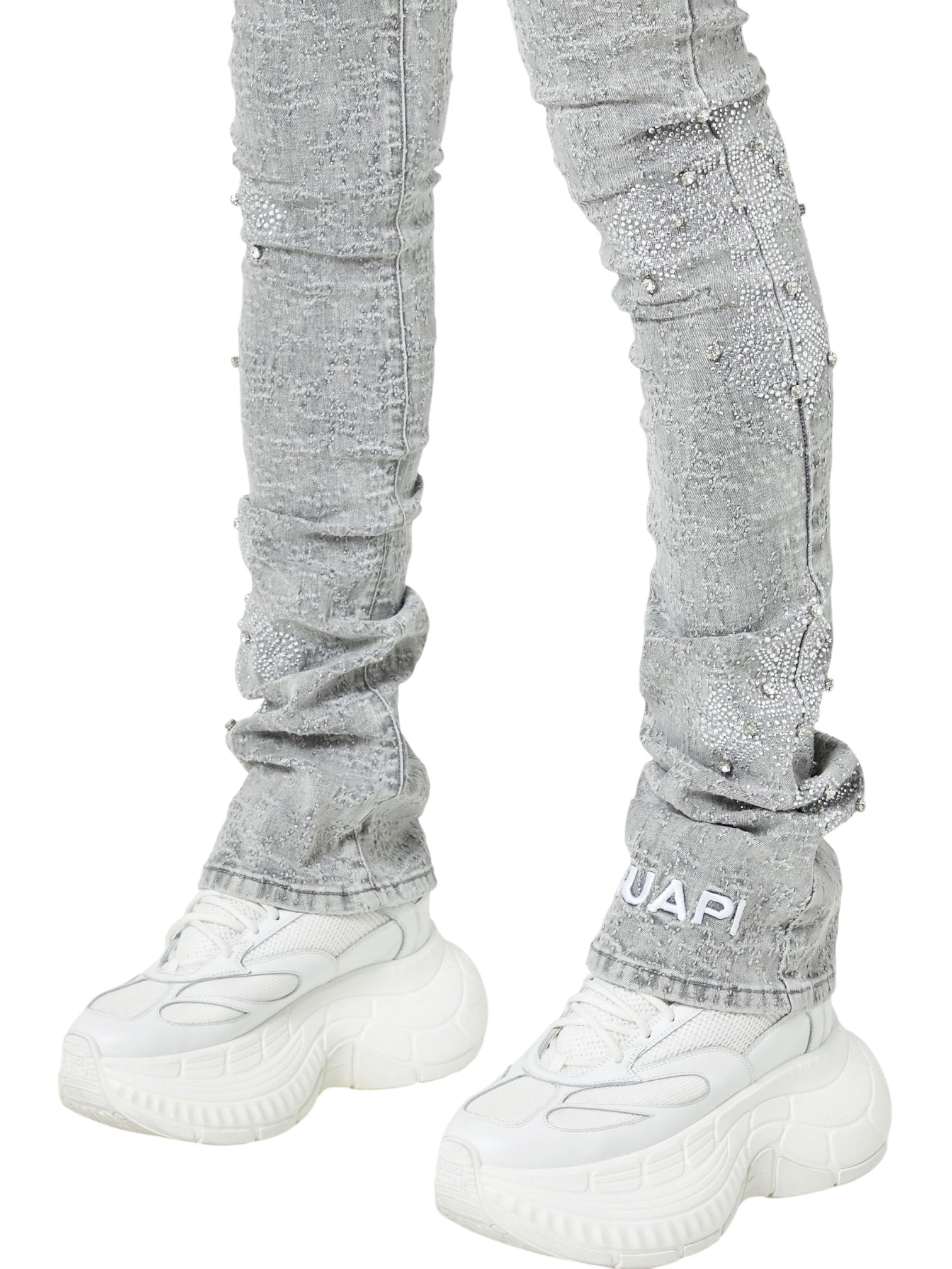 Person wearing GUAPI Smoke Grey Distressed Cross Denim and white chunky sneakers, standing with feet slightly apart. The jeans feature textured details and "GUAPI" branding near the ankle.