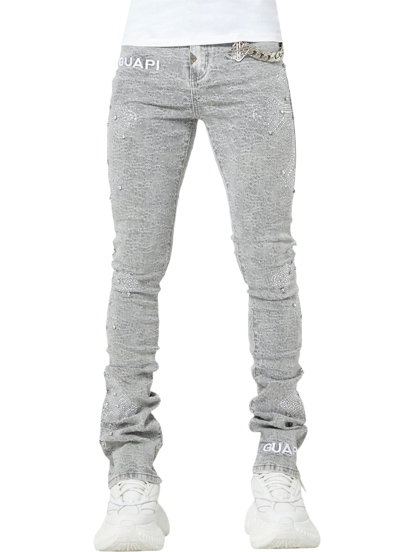 Person wears GUAPI Smoke Grey Distressed Cross Denim with rhinestone cross details, silver chain accent, and "GUAPI" branding, paired with white sneakers. Only the lower body is visible.