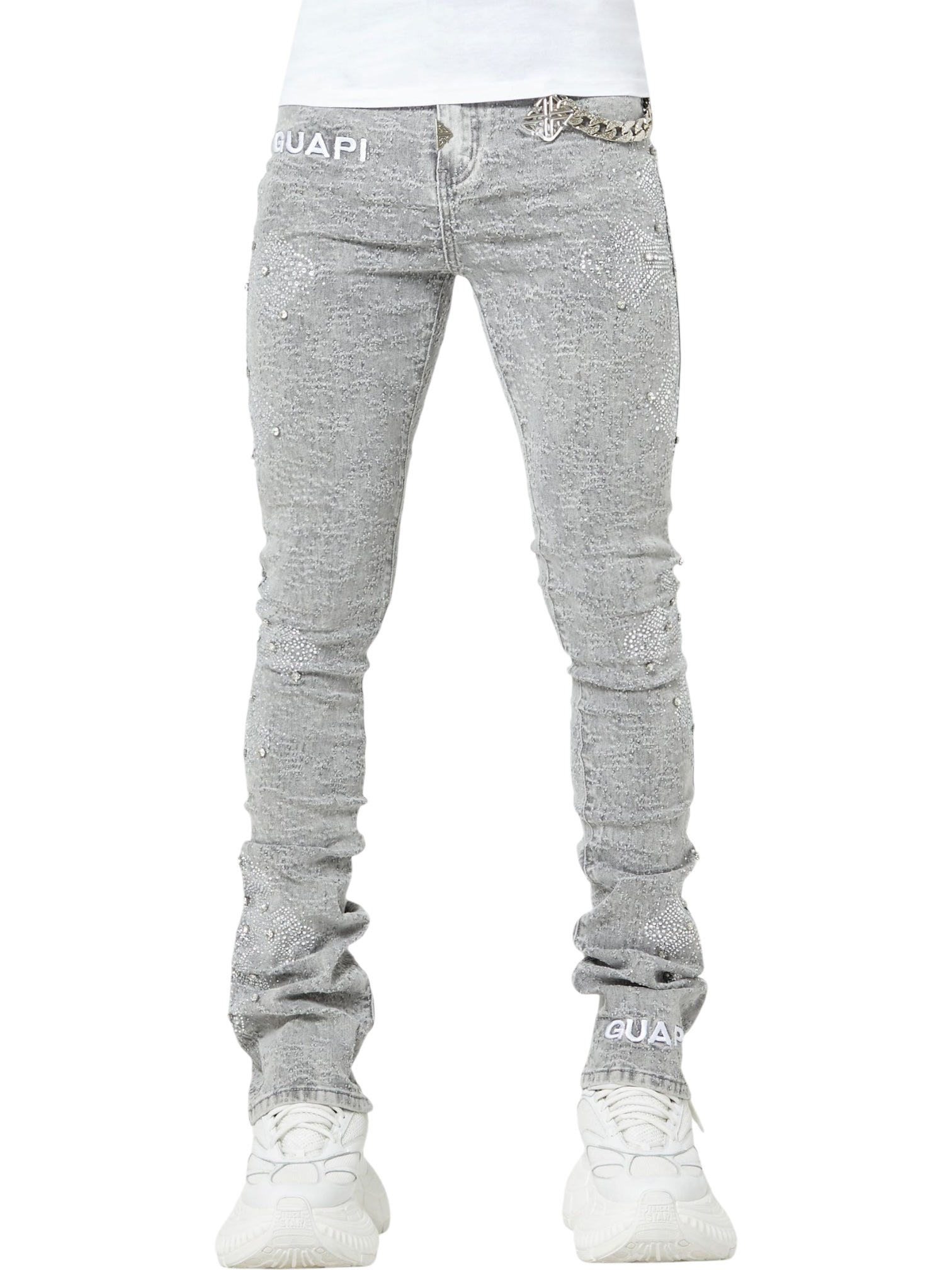 Person wears GUAPI Smoke Grey Distressed Cross Denim with rhinestone cross details, silver chain accent, and "GUAPI" branding, paired with white sneakers. Only the lower body is visible.
