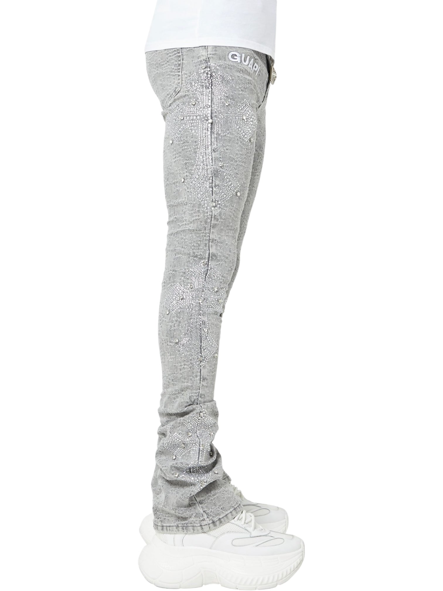 A person in GUAPI Guapi Smoke Grey Distressed Cross Denim jeans and white chunky sneakers, shown in side profile against a white background.