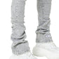 Person wearing Guapi Smoke Grey Distressed Cross Denim by GUAPI, featuring rhinestone cross details and "GU" branding near the ankle, styled with chunky white sneakers. Only lower legs and feet are visible.