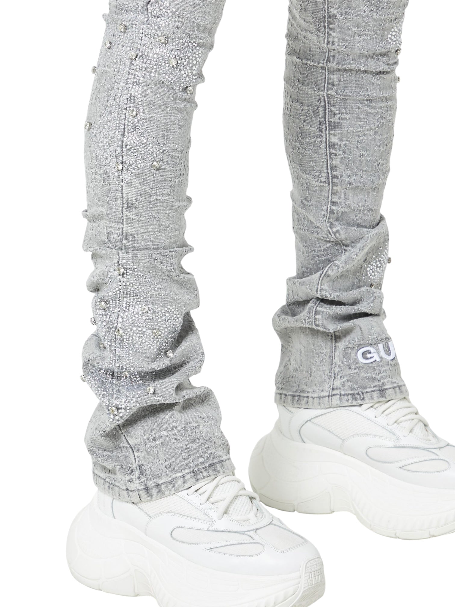 Person wearing Guapi Smoke Grey Distressed Cross Denim by GUAPI, featuring rhinestone cross details and "GU" branding near the ankle, styled with chunky white sneakers. Only lower legs and feet are visible.