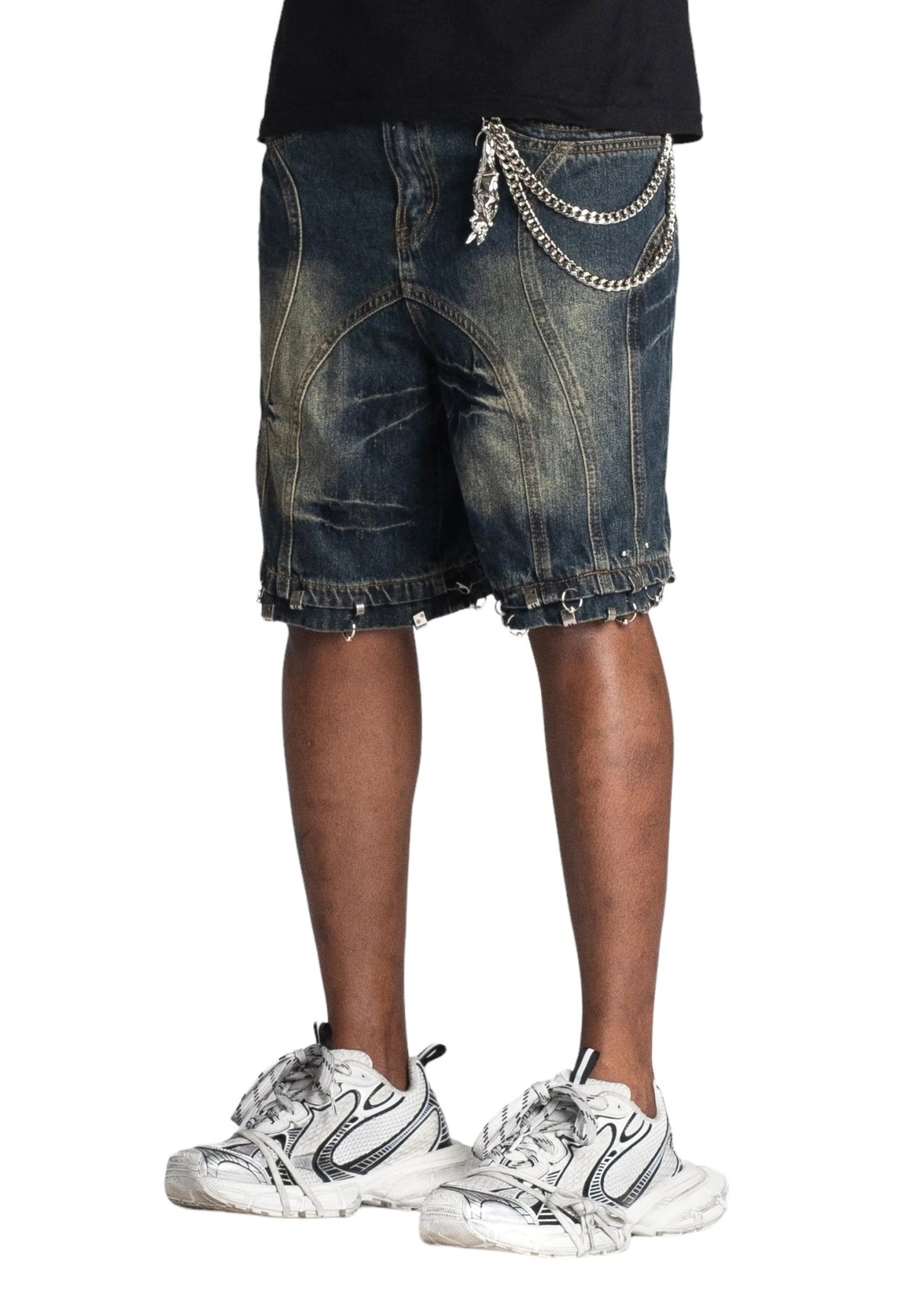 A person wearing GUAPI TEAL BLUE PIERCING SHORTS with chains and white sneakers stands against a white background, showcasing a baggy fit with custom hardware accents.