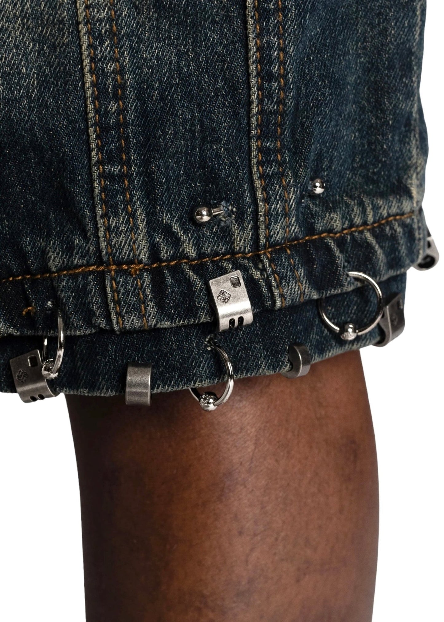 Close-up of a person's leg wearing GUAPI TEAL BLUE PIERCING SHORTS with metal embellishments, including rings and clips, on the bottom hem. These cotton shorts feature a baggy fit, embodying the edgy style of GUAPI.