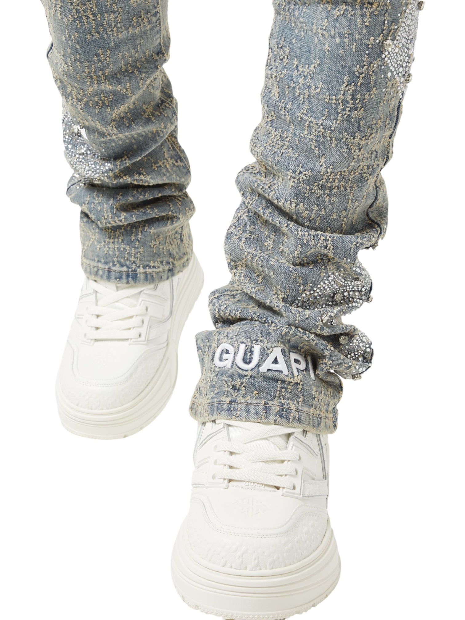 A person wears GUAPI Vintage Blue Distressed Cross Denim with rhinestone details, a "GUAPI" patch on one cuff, and white sneakers.