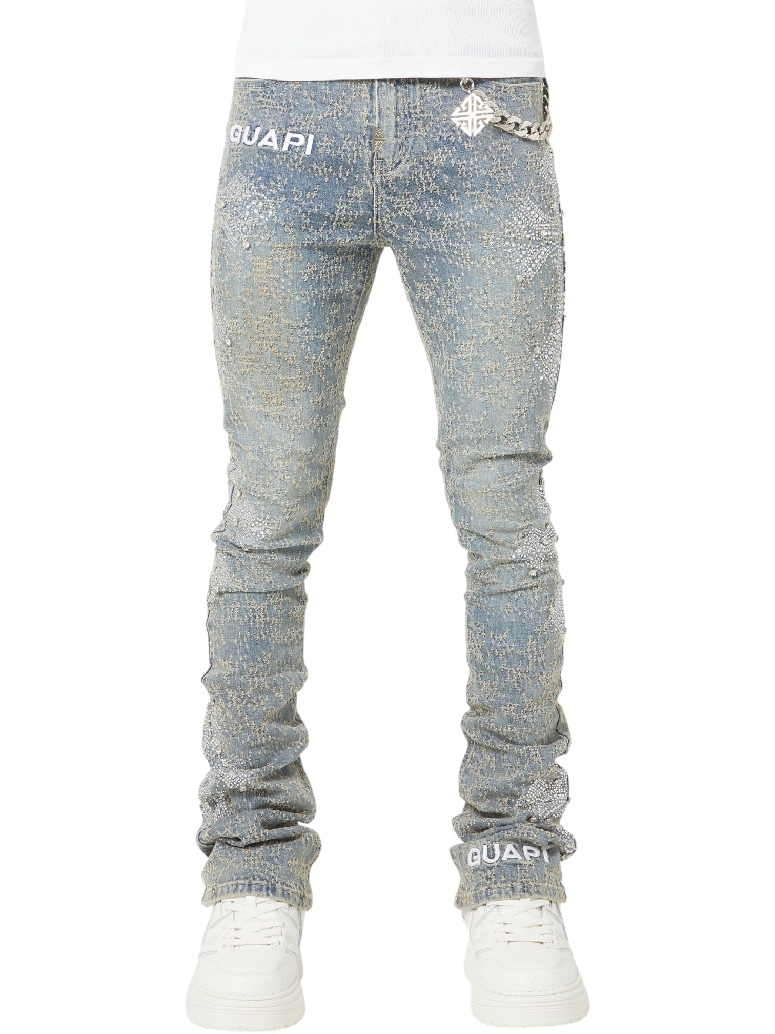 A person wearing GUAPI Vintage Blue Distressed Cross Denim jeans, featuring fitted styling, rhinestone "GUAPI" branding, and white sneakers, shown from waist to feet.