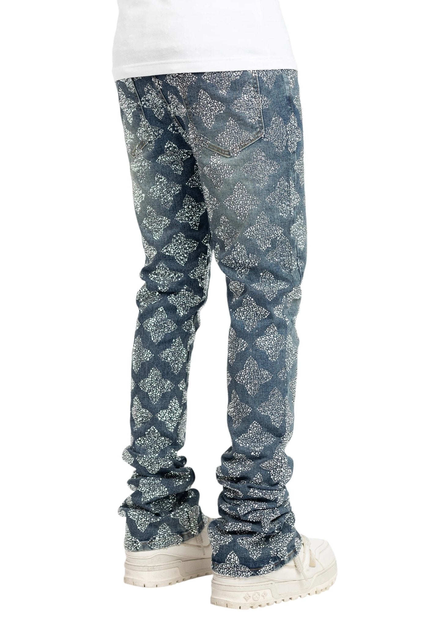 A person confidently stands in slim-fit Guapi Vintage Blue Embellished Denim jeans by GUAPI, showcasing a crystal-studded lattice pattern, with a partially visible white shirt and white sneakers.