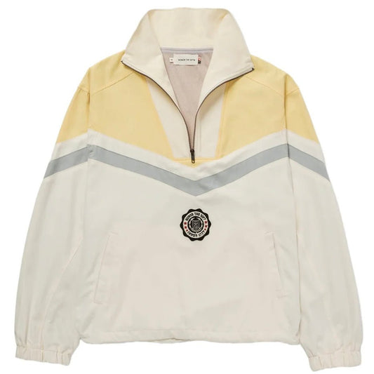 The Honor The Gift A-Spring Brushed Poly Track Anorak in Bone by HONOR THE GIFT is a pullover jacket with a beige and yellow half-zip design, featuring a chevron stripe pattern. Made from durable materials, it boasts a round black emblem on the front, combining style and longevity tailored for the fashion-forward individual.