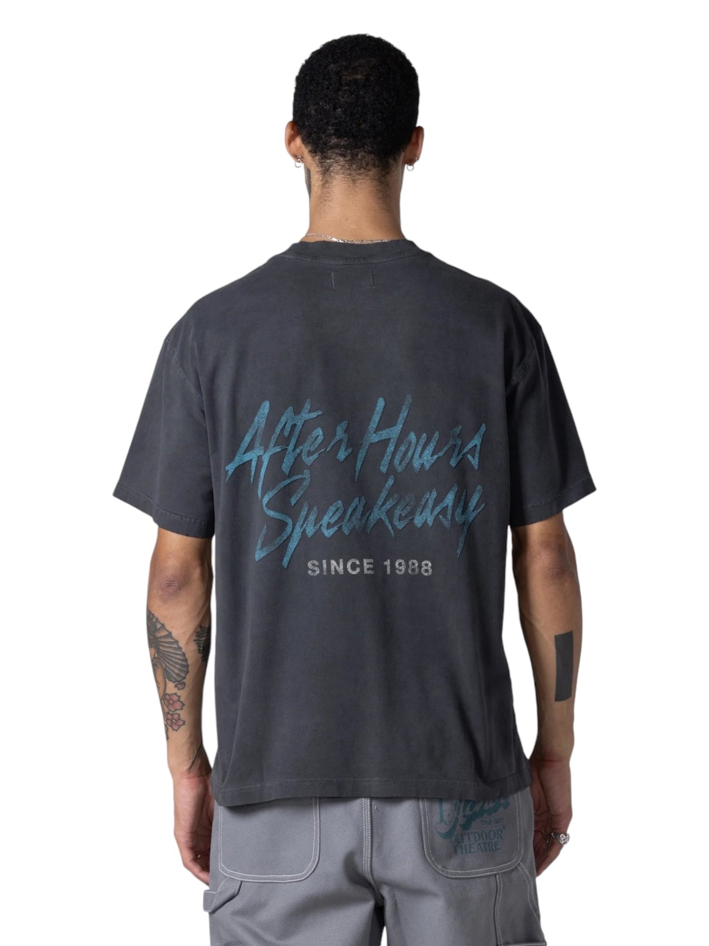 A person stands against a white background wearing the Honor The Gift After Hours T-shirt Black by HONOR THE GIFT, featuring a vintage print, "After Hours Speakeasy Since 1988," made from pigment washed jersey, paired with light pants.