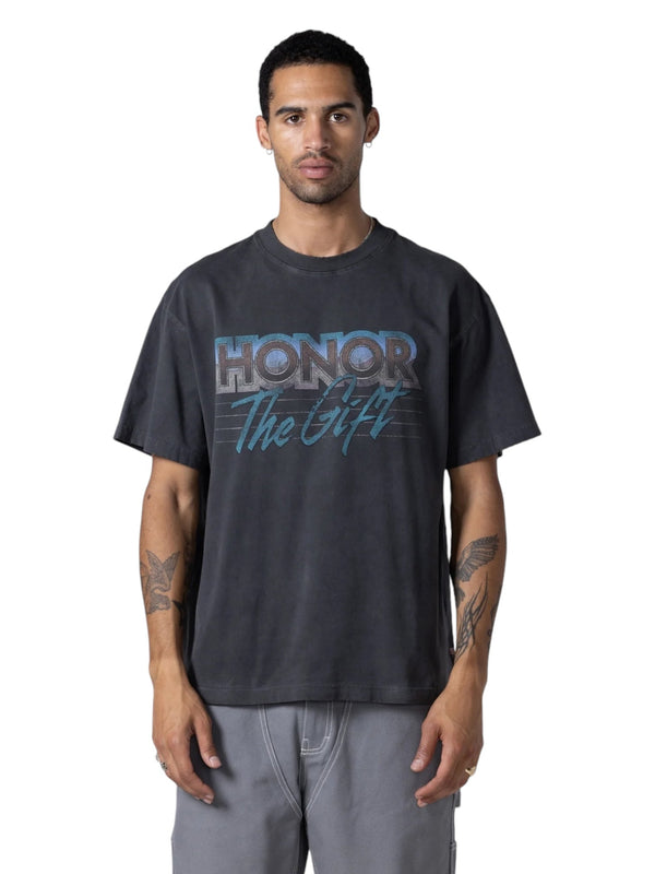 A person in a faded "Honor The Gift After Hours" black T-shirt with a vintage print from HONOR THE GIFT and grey pants presents a classic look; the pigment washed jersey adds uniqueness.