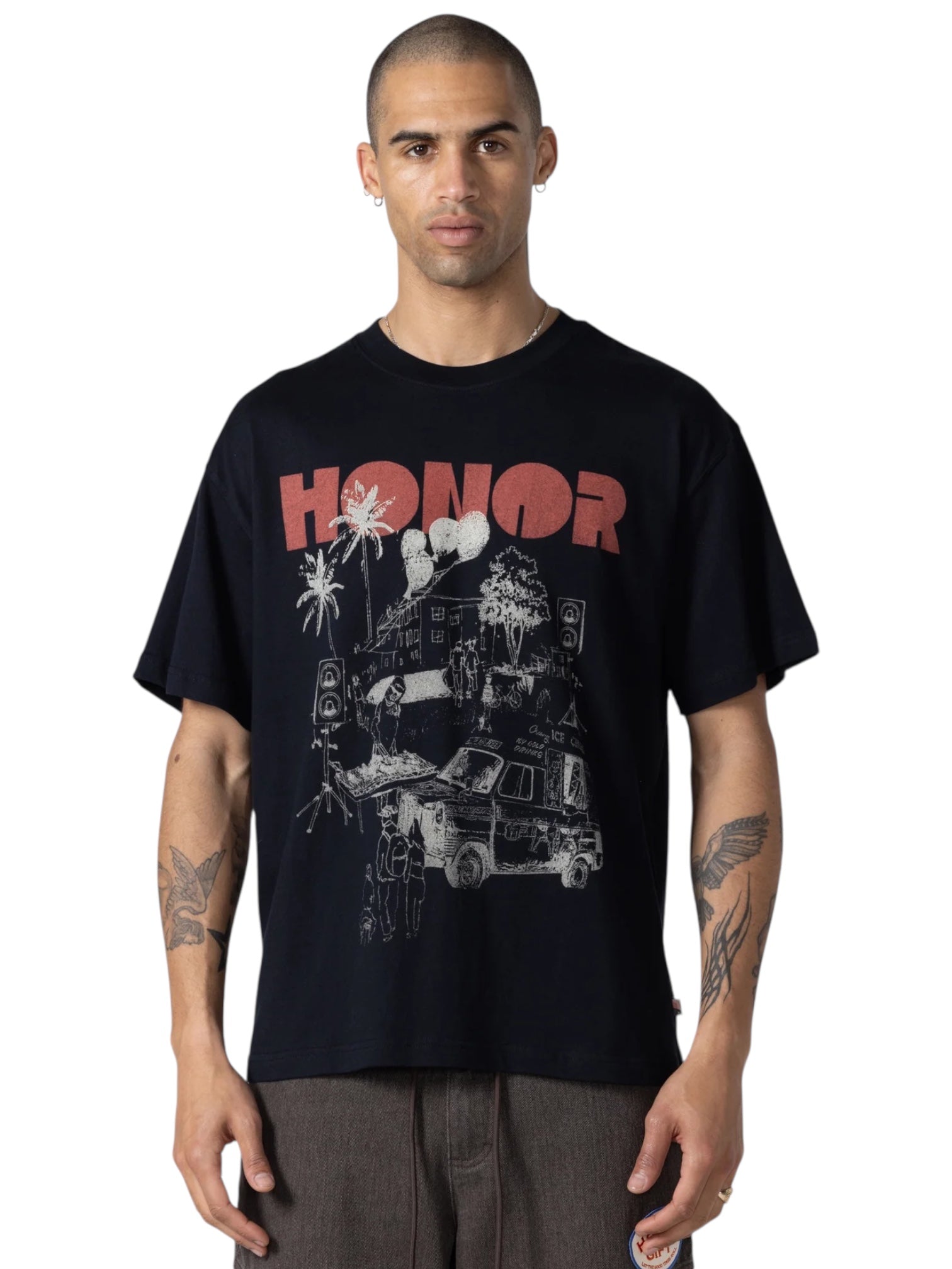 A person stands against a white background wearing the HONOR THE GIFT Block Party T-shirt in black, featuring "HONOR" in red and a house, palm trees, and van graphic. Made from 100% cotton.