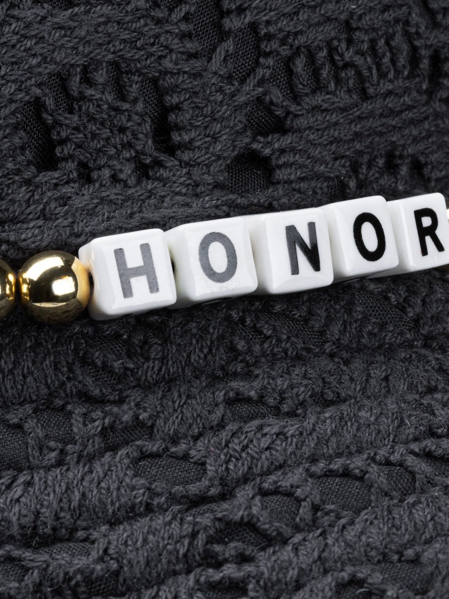 White letter beads spelling "HONOR" and a gold bead are set on black fabric—perfect accents for the HONOR THE GIFT Friendship Crochet Bucket Hat Black by HONOR THE GIFT.