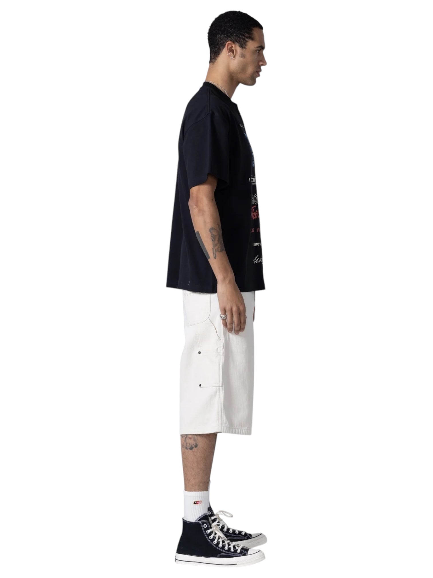 A man stands in profile wearing a black T-shirt, HONOR THE GIFT Gears Carpenter Short Cream with a 13" inseam and 100% cotton, paired with white socks and black high-top sneakers against a plain background.