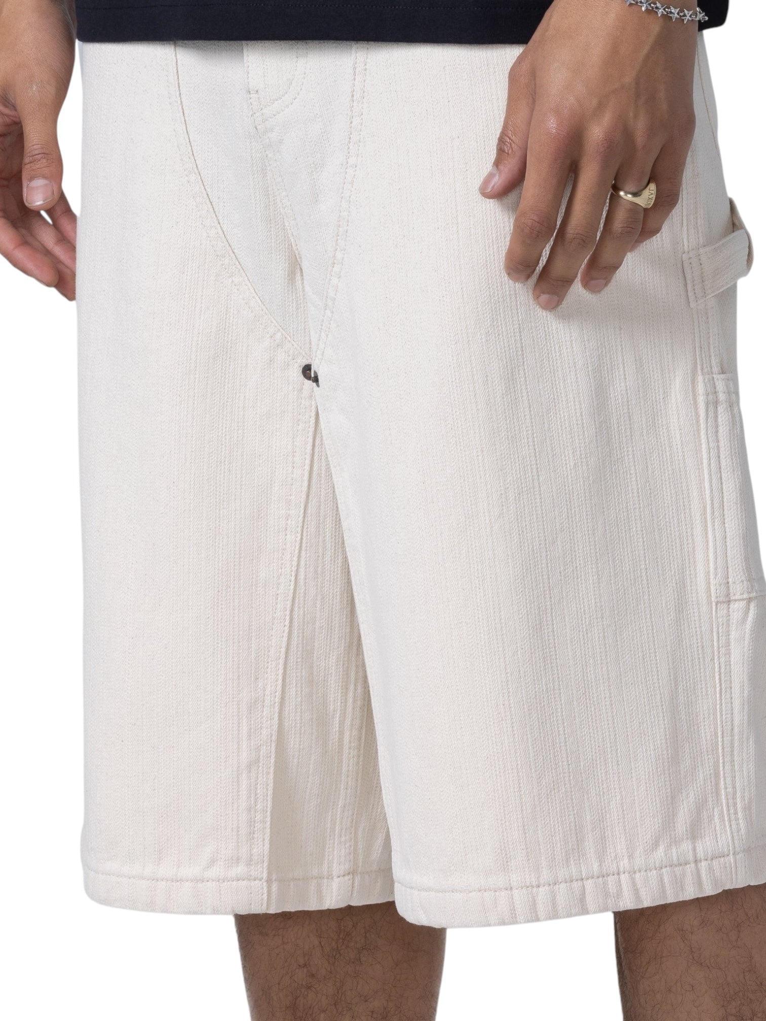 A person wears HONOR THE GIFT’s Honor The Gift Gears Carpenter Short Cream—off-white, 13" inseam, side pockets, 100% cotton—paired with a black shirt, ring, and bracelet for comfort and style.