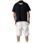 A person stands facing away, wearing a black tee with a back graphic and HONOR THE GIFT Gears Carpenter Short Cream, featuring 13" inseam white twill made from 100% cotton, paired with white socks and black high-top sneakers.