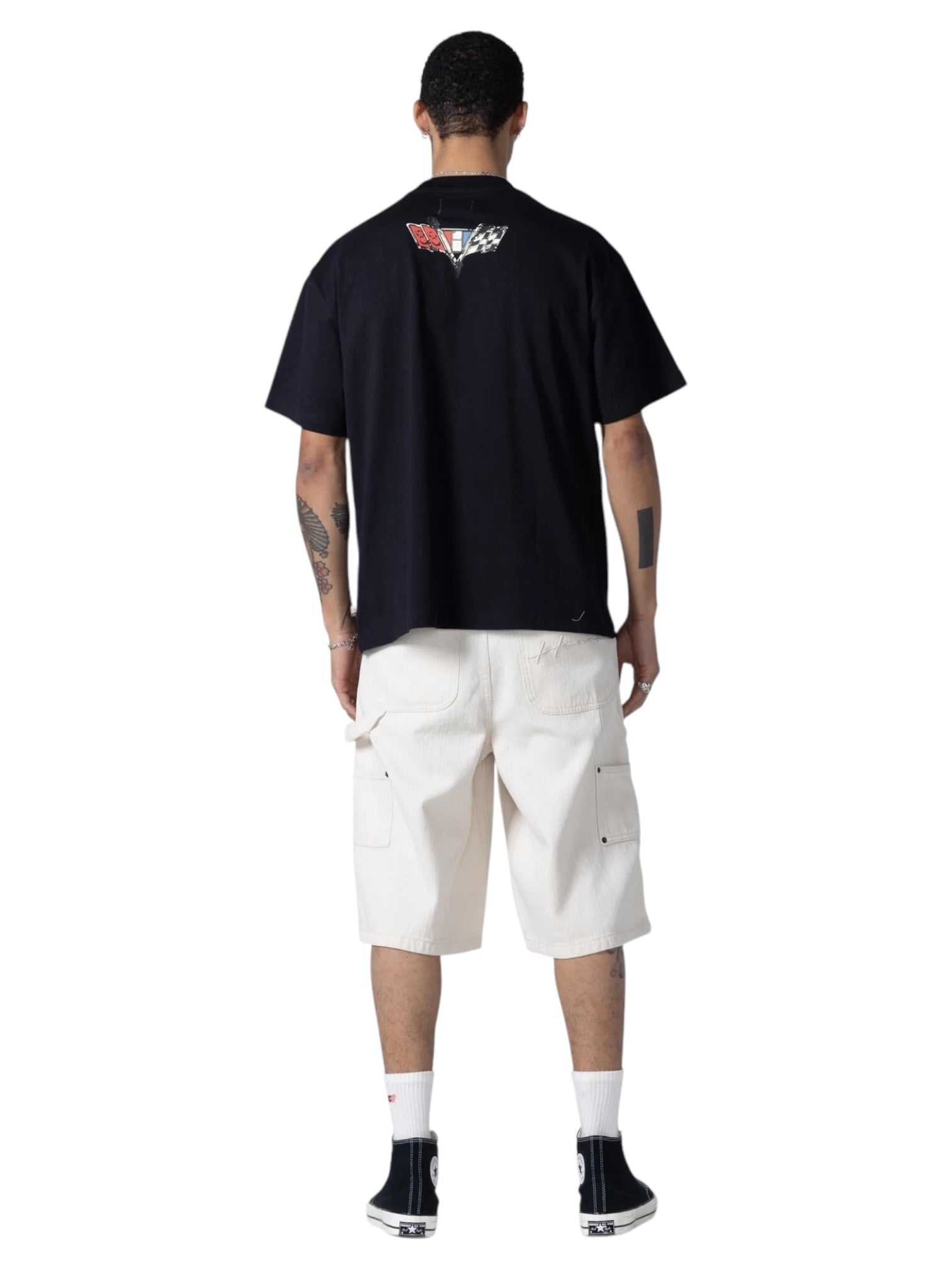 A person stands facing away, wearing a black tee with a back graphic and HONOR THE GIFT Gears Carpenter Short Cream, featuring 13" inseam white twill made from 100% cotton, paired with white socks and black high-top sneakers.