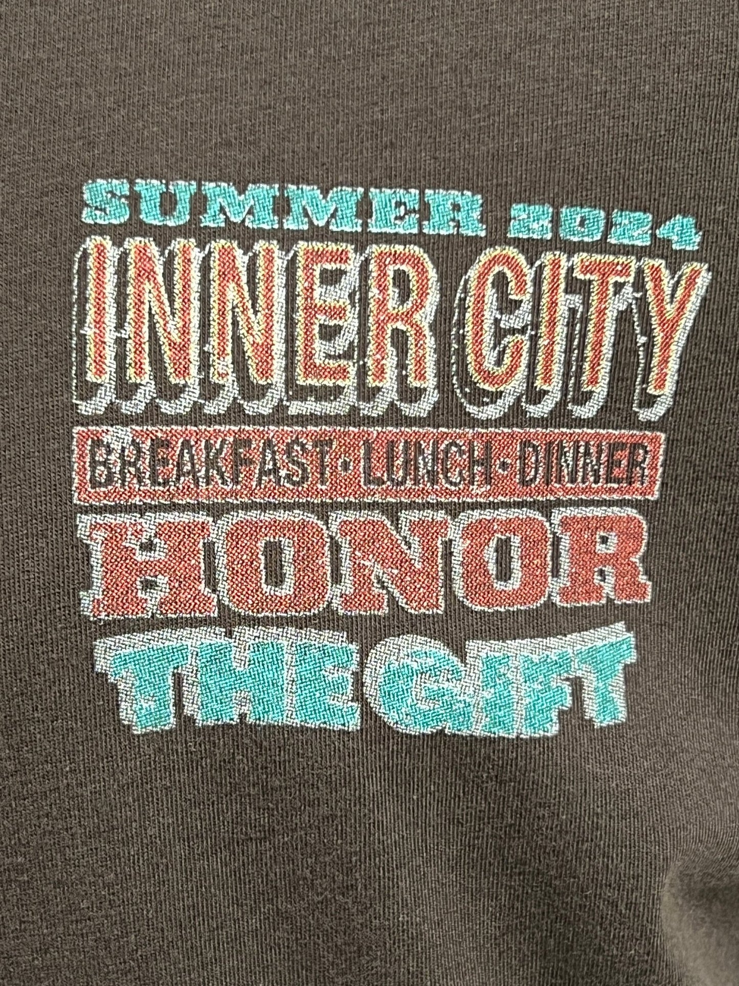 The HONOR THE GIFT HTG BURGERS SS TEE BLACK is a vintage-style, 100% cotton pocket T-shirt featuring the text "Summer 2024 Inner City - Breakfast, Lunch, Dinner - Honor The Gift," printed in different colors and fonts. Garment dyed for a soft feel, it's perfect for any casual outing.