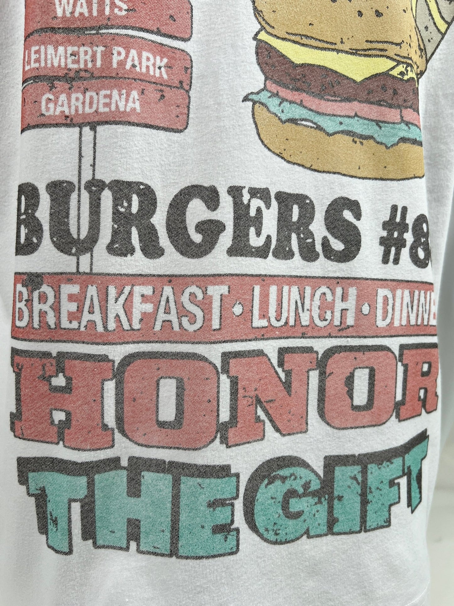 A close-up of a HONOR THE GIFT HTG BURGERS SS TEE WHITE with text and graphics. The text reads, "Watts, Leimert Park, Gardena, Burgers #8, Breakfast, Lunch, Dinner, Honor the Gift." It features a burger illustration.