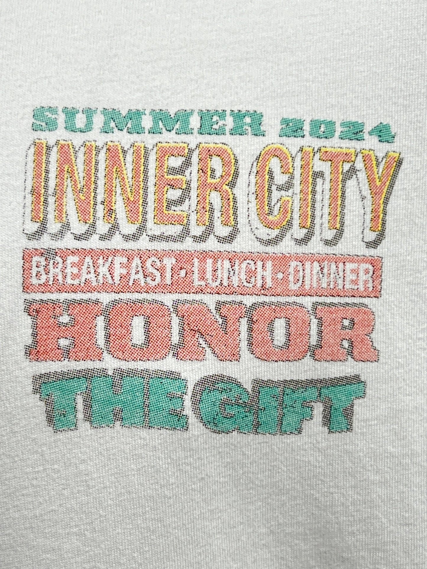 Text on a HONOR THE GIFT HTG BURGERS SS TEE WHITE reads "SUMMER 2024 INNER CITY BREAKFAST LUNCH DINNER HONOR THE GIFT" in colorful, stylized fonts with a touch of retro charm.