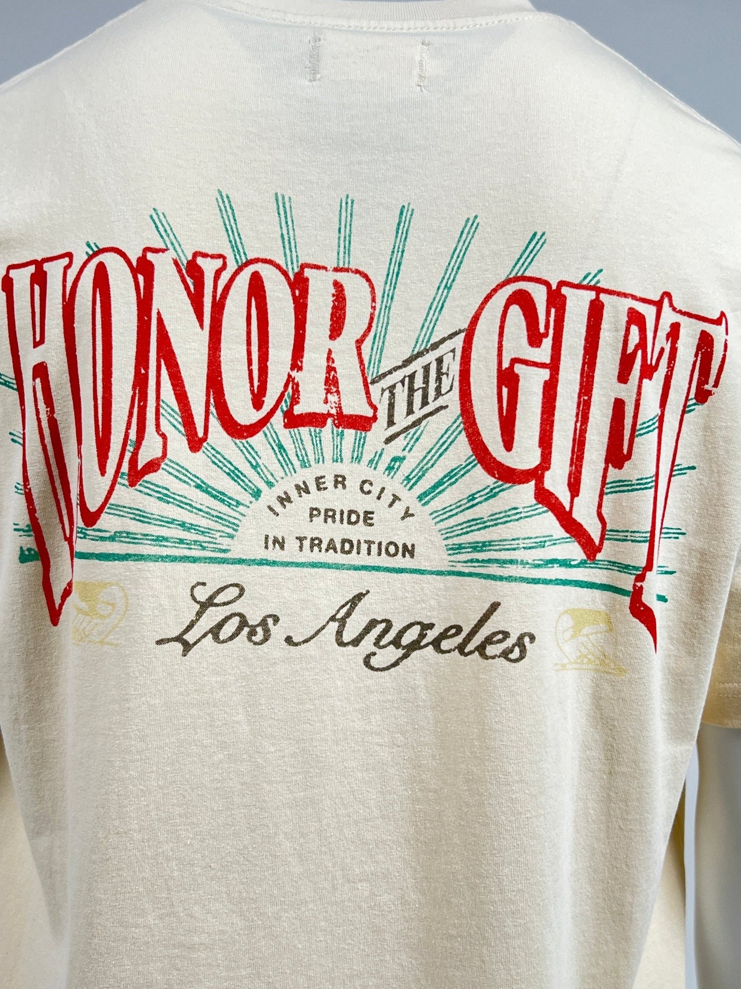 The HONOR THE GIFT HTG CIGAR LABEL SS TEE BONE is a white 100% cotton T-shirt featuring the text "HONOR THE GIFT" in red and "INNER CITY PRIDE IN TRADITION Los Angeles" in green and black on the back, styled to resemble a vintage cigar label.