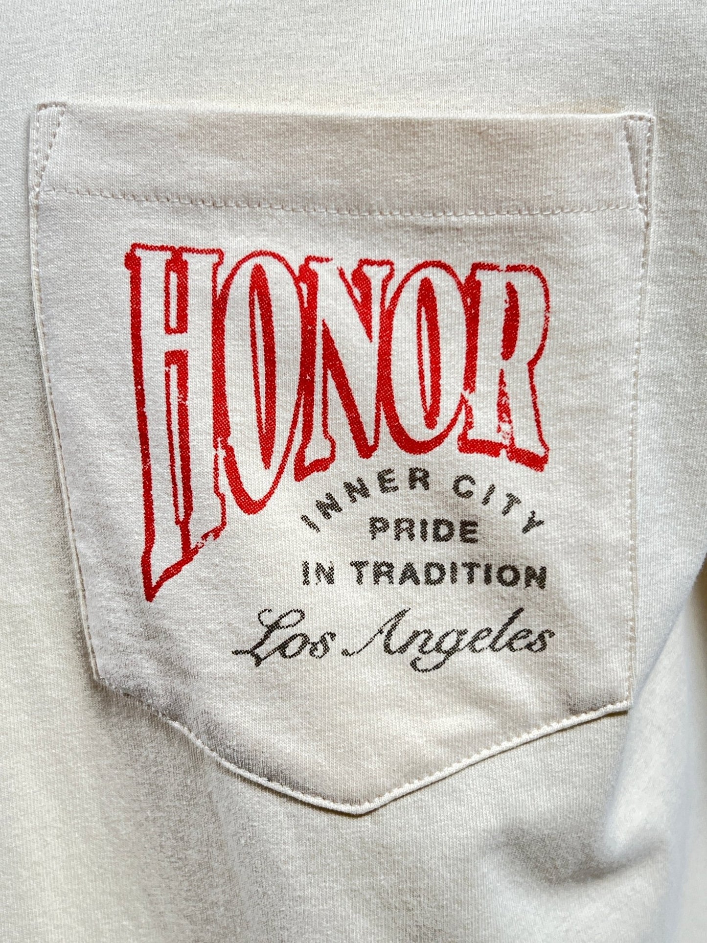 Close-up of the HONOR THE GIFT HTG CIGAR LABEL SS TEE BONE's white pocket, featuring the word "HONOR" in large red letters followed by "INNER CITY PRIDE IN TRADITION Los Angeles" in smaller black letters. This 100% cotton shirt, offered by HONOR THE GIFT, provides lightweight comfort for everyday wear.