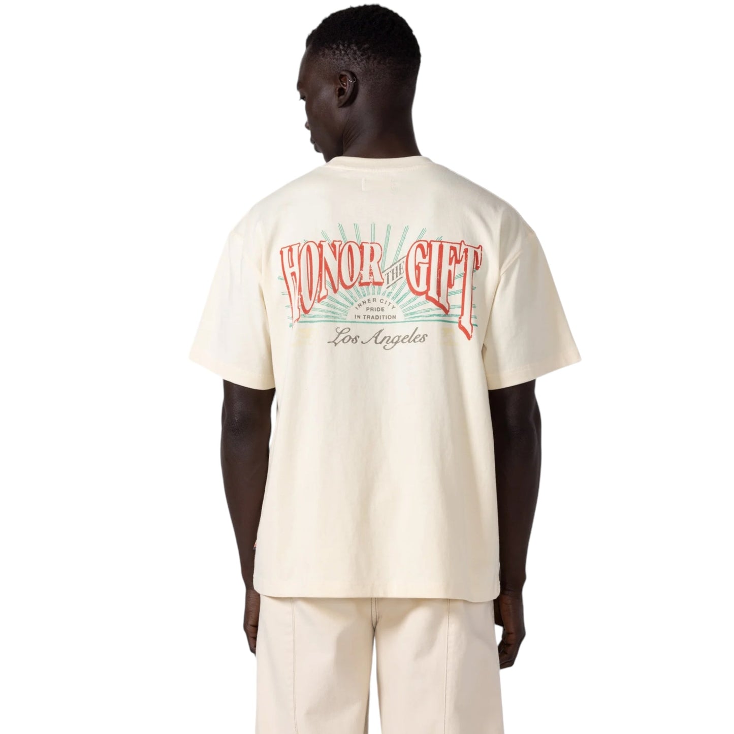 A person wears the Honor The Gift Htg Cigar Label Ss Tee in bone, featuring "HONOR THE GIFT" and "Los Angeles" elegantly printed on the back. This vintage-style T-shirt is crafted from 100% cotton and pairs seamlessly with cream pants.