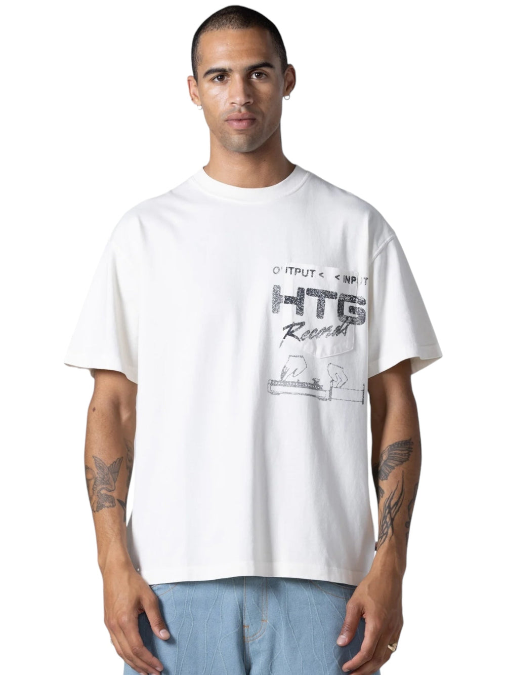 A person in a loose HONOR THE GIFT HTG RECORDS TEE WHITE, showcasing a distressed custom artwork graphic, stands facing forward. They have short hair and visible tattoos on their forearms.