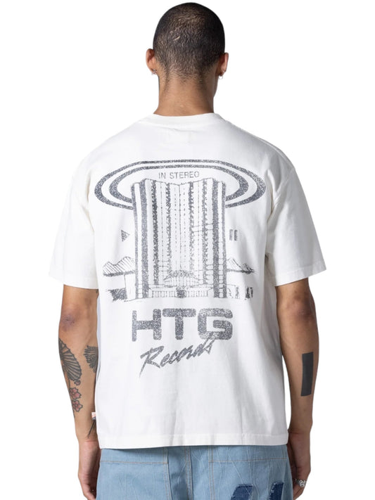 A person wearing the HONOR THE GIFT HTG RECORDS TEE WHITE, made of 100% cotton, with a graphic design featuring the words "In Stereo" and "HTG Record" on the back, shown from behind. The person has tattoos on both arms, is wearing blue jeans, and the shirt showcases distressed custom artwork for a vintage wash effect.