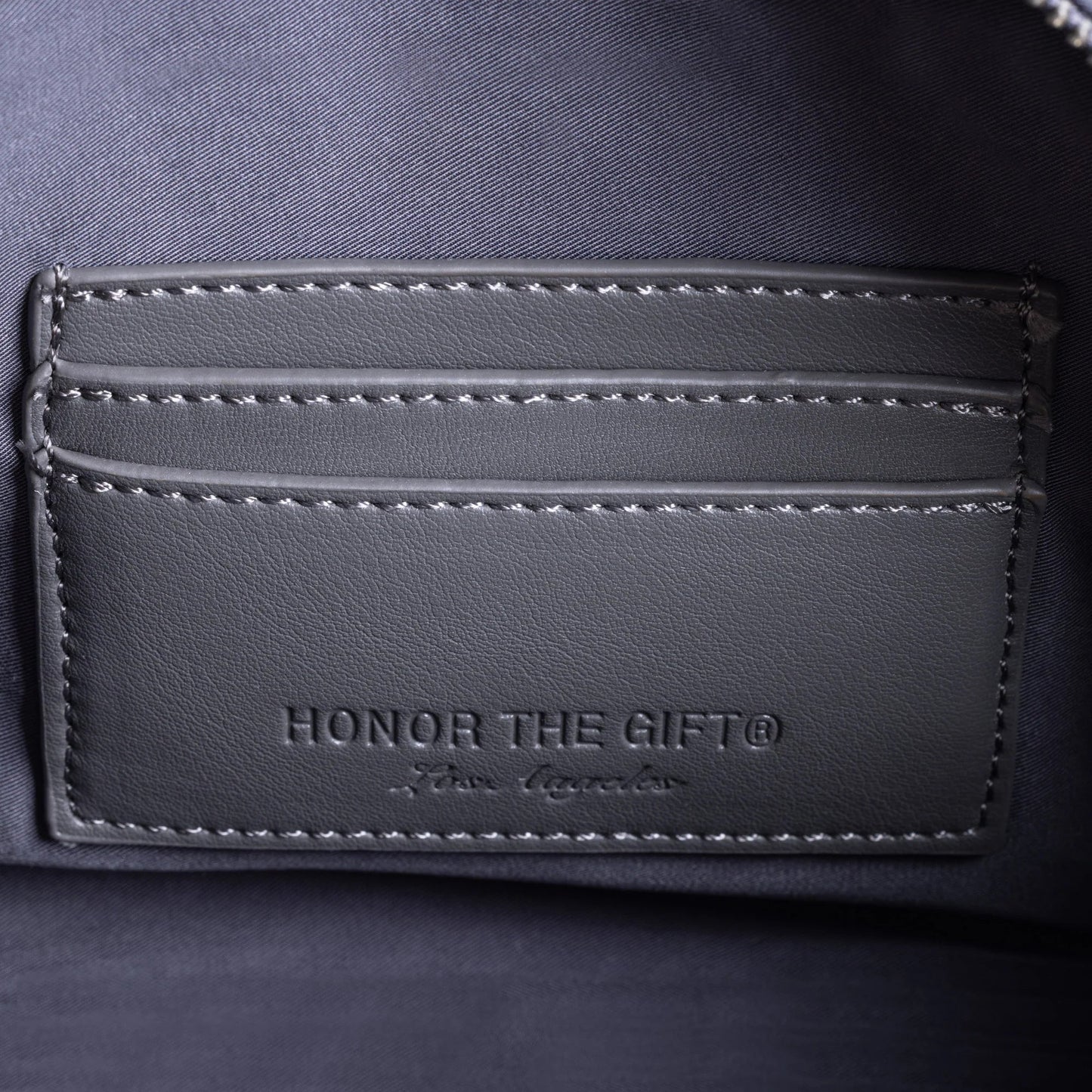 Close-up of a dark grey side bag featuring a cardholder section and an "H" logo closure. The text "HONOR THE GIFT®" and "From Signature" is embossed on the faux leather.