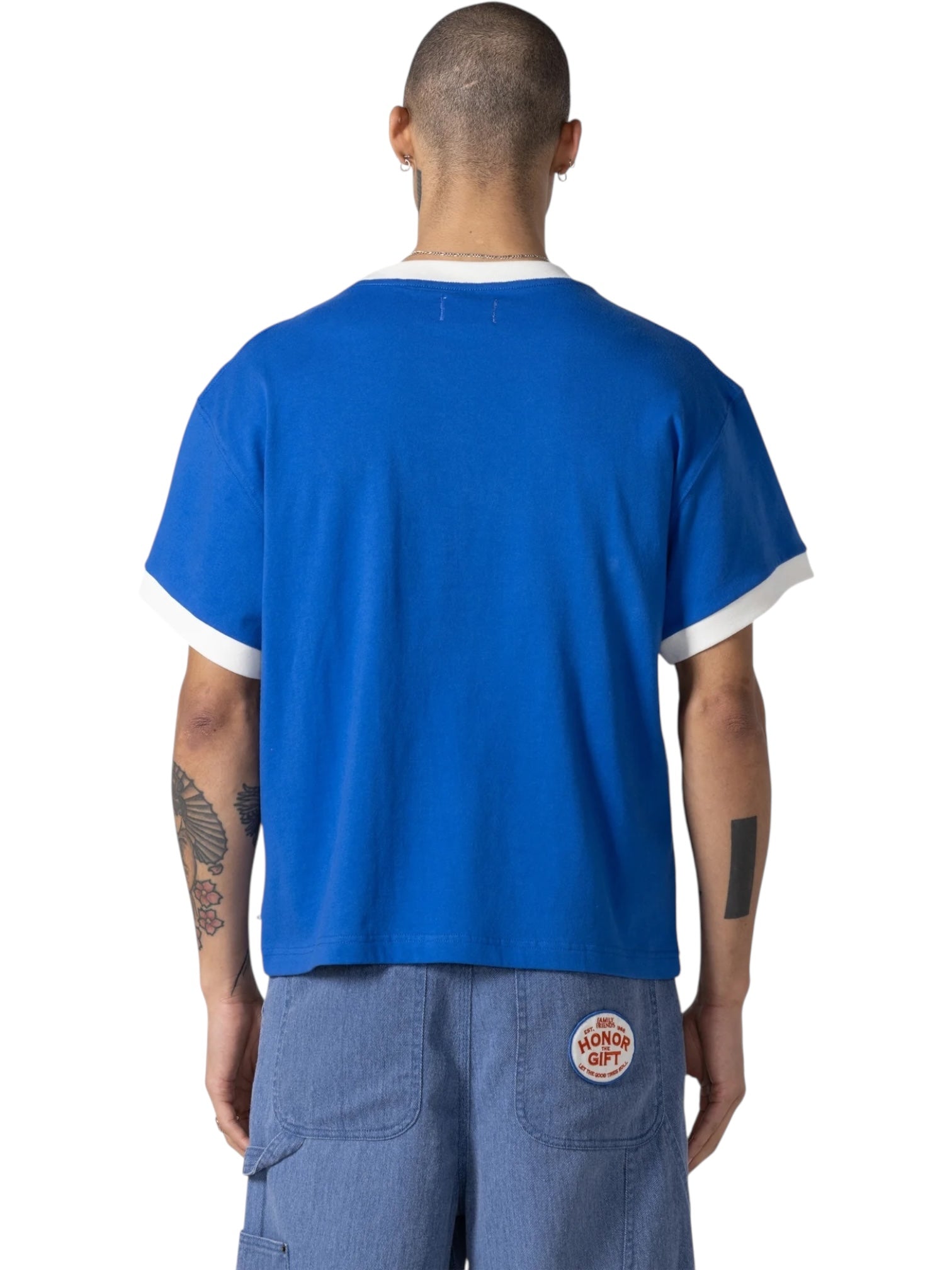 A person with short hair faces away, wearing the HONOR THE GIFT Multi Box Tee Blue with white trim and blue shorts featuring a circular patch on the pocket. Tattoos are visible on both arms.
