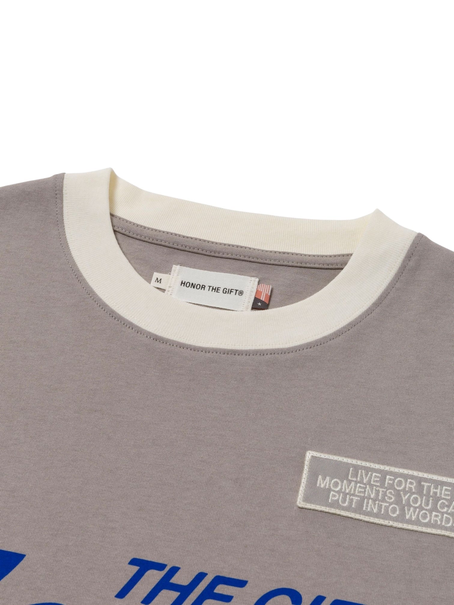 Close-up of the HONOR THE GIFT Multi Box Tee Stone, a gray heavyweight cotton t-shirt with a white collar, visible tags, and a patch reading "LIVE FOR THE MOMENTS YOU CAN’T PUT INTO WORDS." Blue text adds a touch of style to this durable piece.