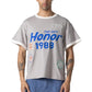 A person wears the HONOR THE GIFT Multi Box Tee Stone, a structured gray heavyweight cotton t-shirt with bold blue "The Gift Honor 1988" text and patches, styled with blue patch pants for durable, standout wear.
