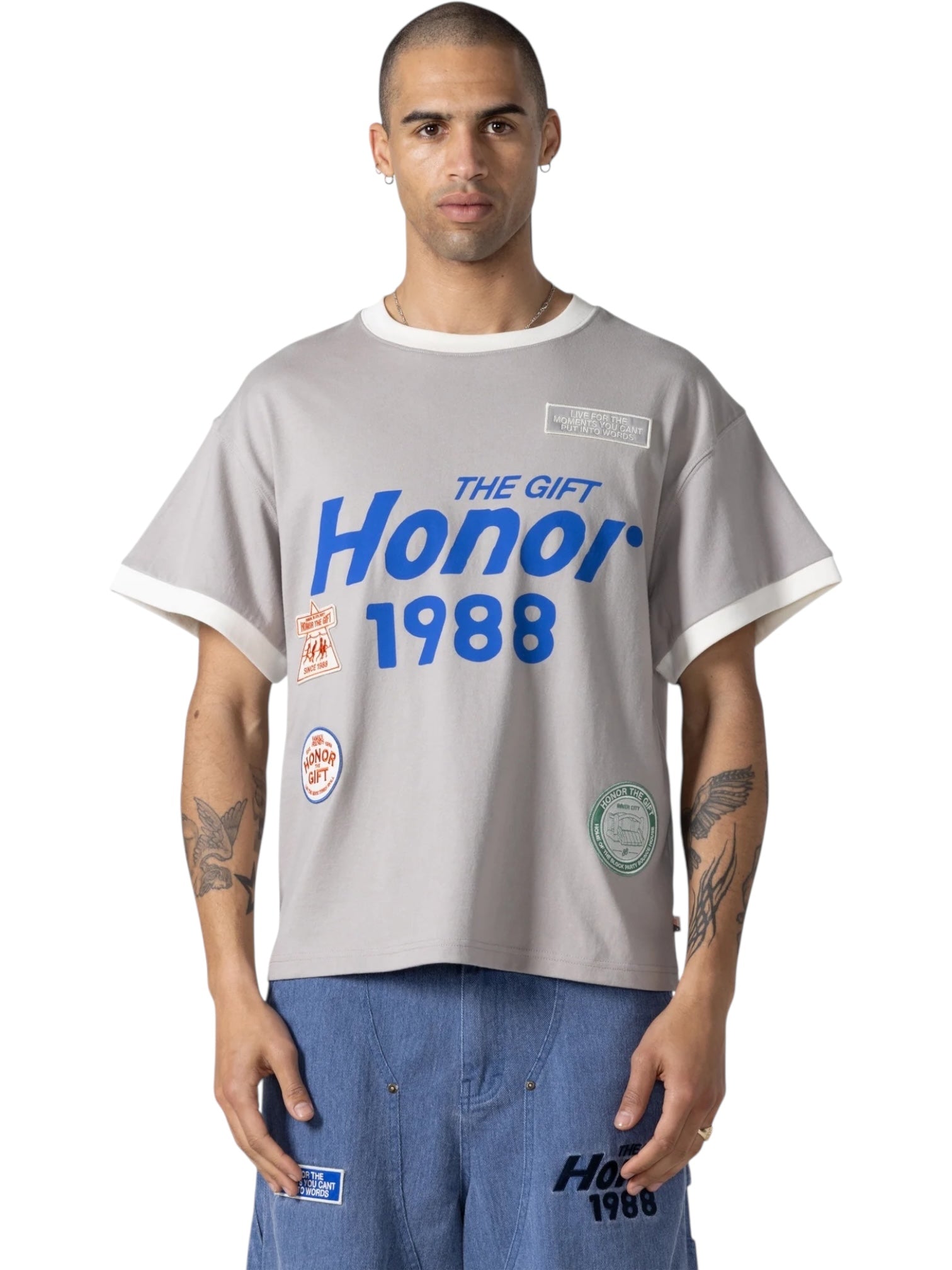 A person wears the HONOR THE GIFT Multi Box Tee Stone, a structured gray heavyweight cotton t-shirt with bold blue "The Gift Honor 1988" text and patches, styled with blue patch pants for durable, standout wear.