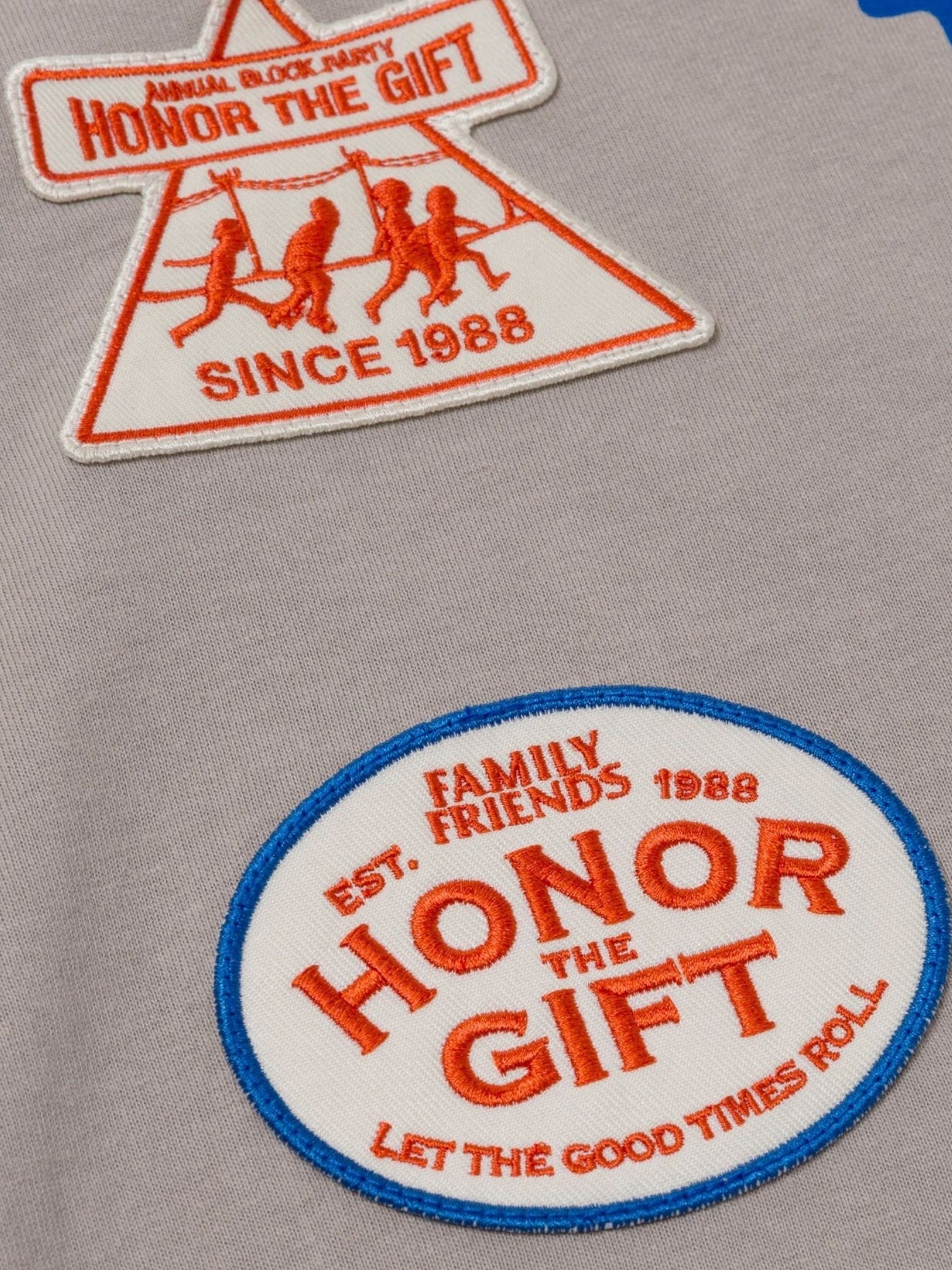 The Honor The Gift Multi Box Tee Stone features two embroidered patches—one triangular with "HONOR THE GIFT SINCE 1988" and human figures, and one oval with "HONOR THE GIFT" in red and blue on heavyweight cotton for lasting wear.