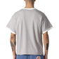 A person with short hair and tattoos is seen from the back wearing the HONOR THE GIFT Multi Box Tee Stone, a light gray heavyweight cotton t-shirt, paired with blue shorts featuring a red "Honor The Gift" patch.