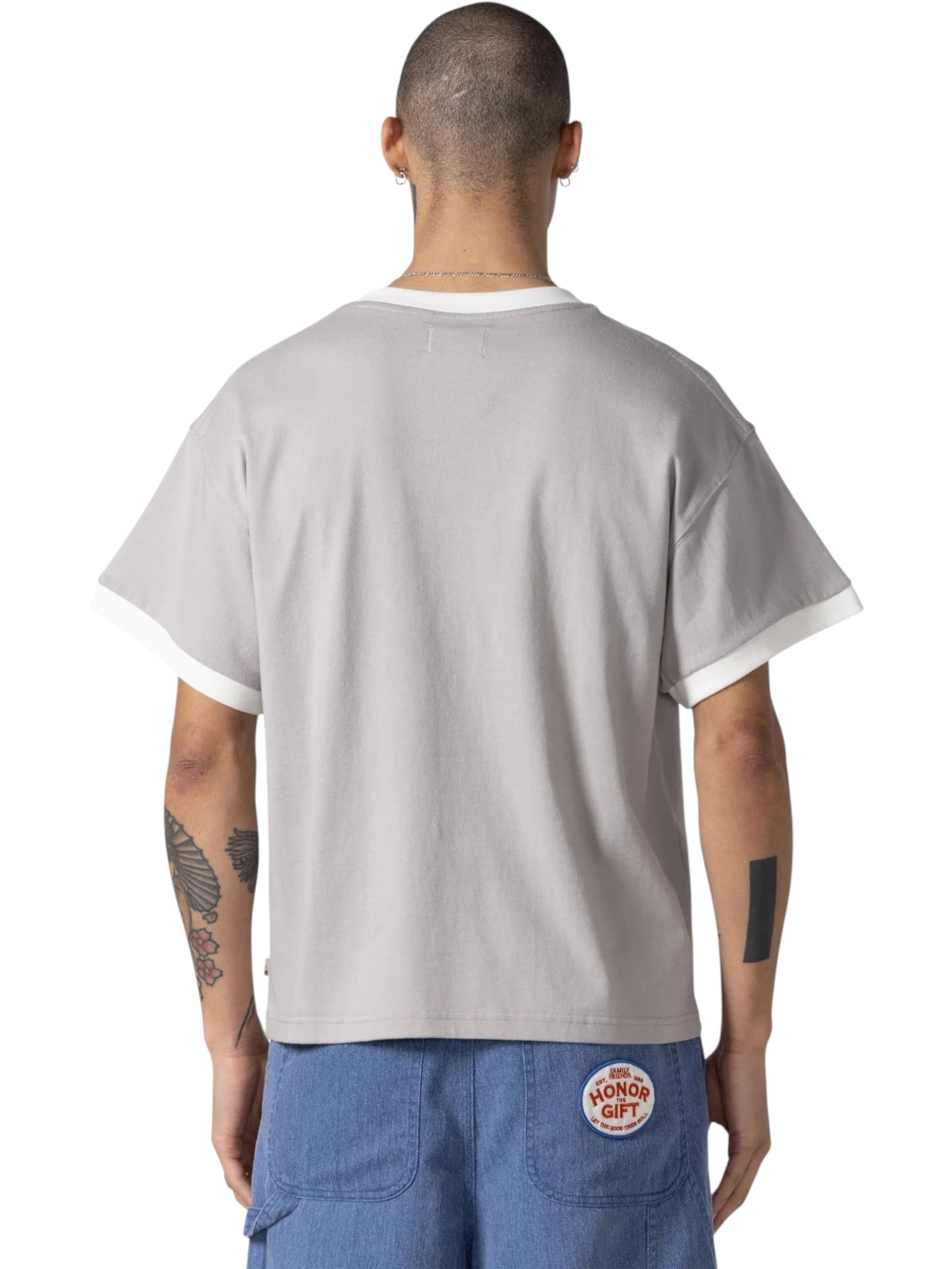 A person with short hair and tattoos is seen from the back wearing the HONOR THE GIFT Multi Box Tee Stone, a light gray heavyweight cotton t-shirt, paired with blue shorts featuring a red "Honor The Gift" patch.