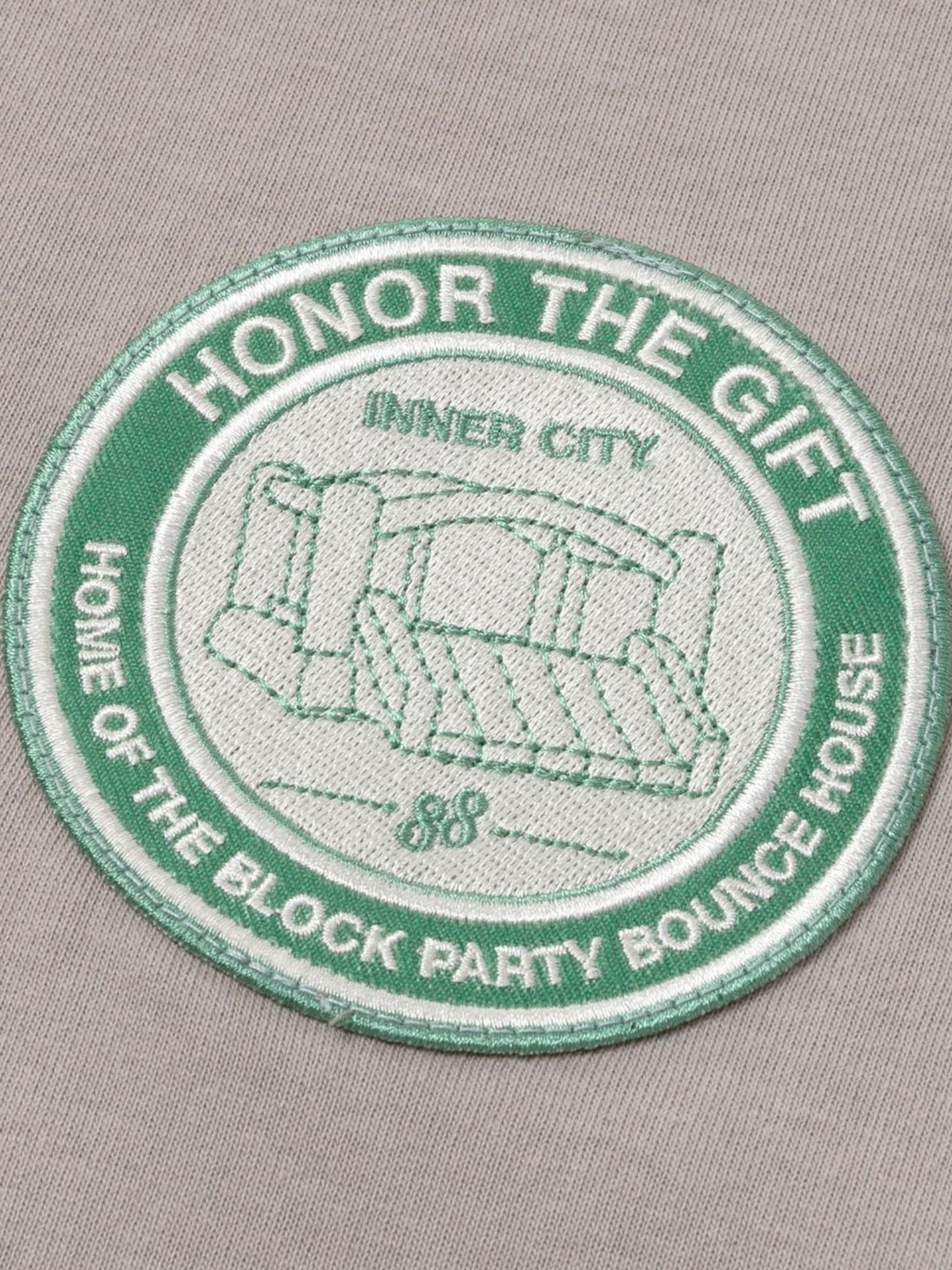 The HONOR THE GIFT Multi Box Tee Stone features a heavyweight cotton build and a patch with green and white "HONOR THE GIFT," "INNER CITY," "HOME OF THE BLOCK PARTY BOUNCE HOUSE" embroidery plus a playful bounce house illustration.