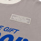 A close-up of the HONOR THE GIFT Honor The Gift Multi Box Tee Stone, a grey and white heavyweight cotton t-shirt featuring a patch with "LIVE FOR THE MOMENTS YOU CAN'T PUT INTO WORDS" and partially visible blue text below.