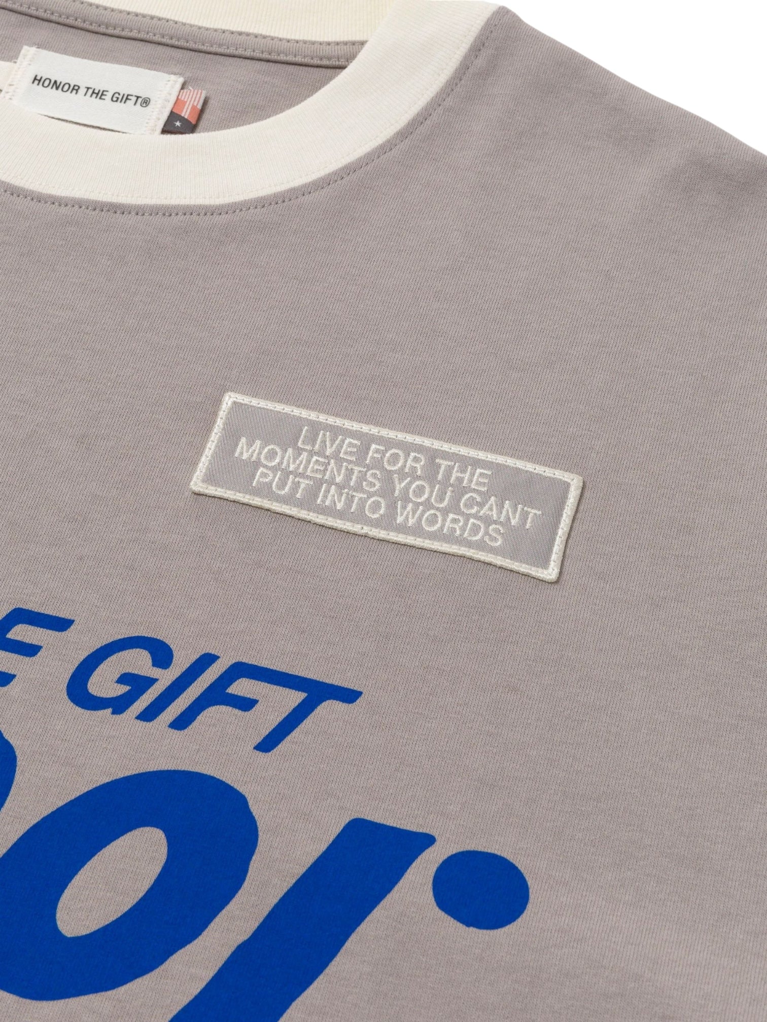A close-up of the HONOR THE GIFT Honor The Gift Multi Box Tee Stone, a grey and white heavyweight cotton t-shirt featuring a patch with "LIVE FOR THE MOMENTS YOU CAN'T PUT INTO WORDS" and partially visible blue text below.