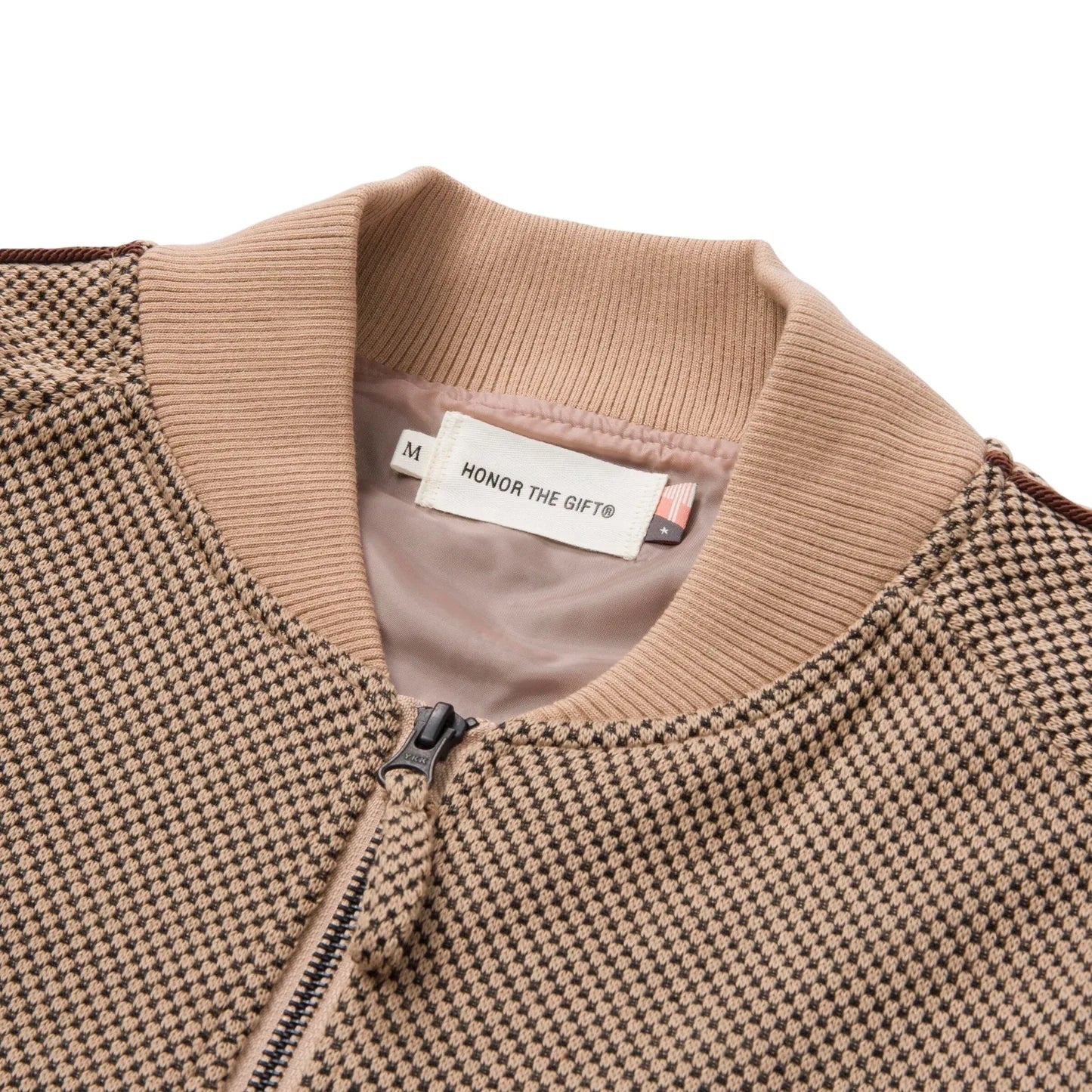 A close-up of the Honor The Gift Novelty Knit Track Jacket Brown reveals its checkered design with a ribbed collar and zip front closure. Inside, a label reads "HONOR THE GIFT®." Flat embroidery enhances the textured fabric, which showcases a pattern of beige and dark brown squares.