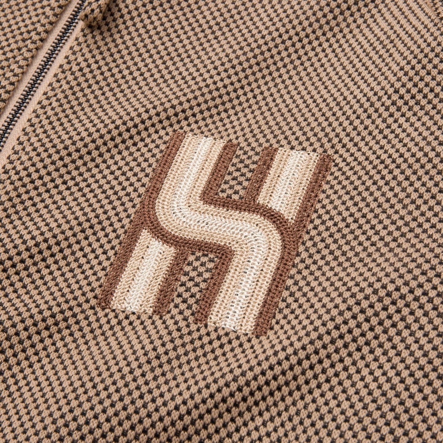 Close-up of a fabric from the HONOR THE GIFT Novelty Knit Track Jacket in Brown, featuring a brown and beige checkered design with a geometric "H" pattern stitched in white and brown threads, reminiscent of intricate flat embroidery.