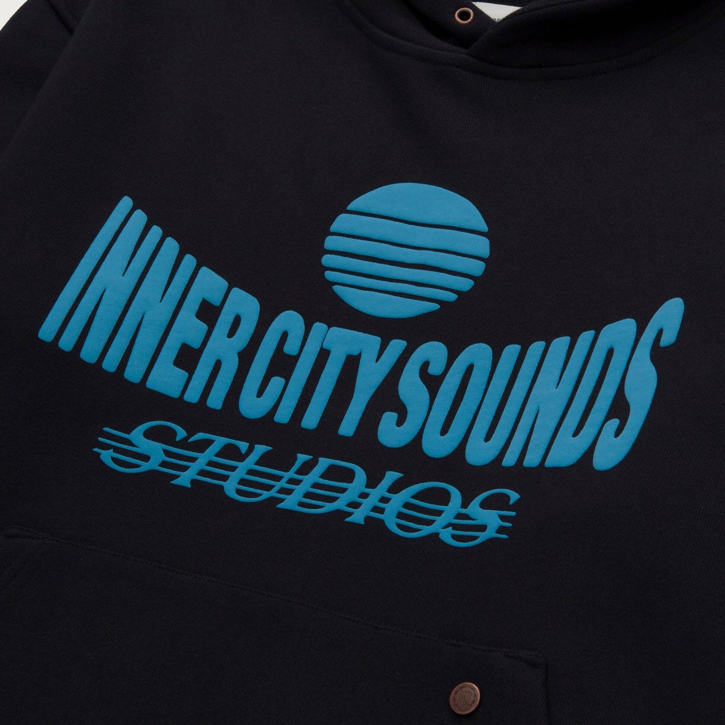 The Honor The Gift Studio Hoodie Black by HONOR THE GIFT features a heavyweight Terry material in black, adorned with a light blue puff print of "INNER CITY SOUNDS STUDIOS" text and a stylized sun graphic.