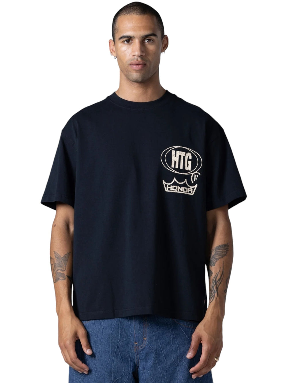 A man wearing the "HONOR THE GIFT STUDIO TEE BLACK" by HONOR THE GIFT, which features "HTG" and "Honda" logos, stands against a plain background. He has short hair and visible tattoos on his arms, with the distressed artwork on the shirt adding an edgy touch to his look.