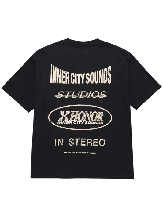 The HONOR THE GIFT STUDIO TEE BLACK from HONOR THE GIFT is a black t-shirt adorned with various white text designs on the back, including "INNER CITY SOUNDS," "STUDIOS," "X HONOR," and "IN STEREO." Made from heavyweight cotton jersey, this piece sports a custom studio print for a distinctive appearance.