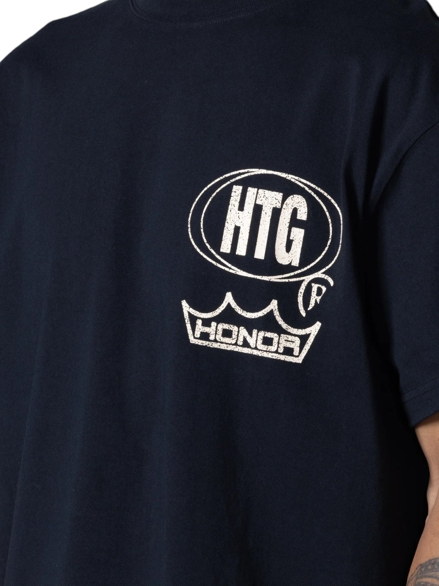 Close-up of the back of an HONOR THE GIFT STUDIO TEE BLACK by HONOR THE GIFT, featuring white printed logos that read "HTG" above a crown symbol and "HONOR." Made from heavyweight cotton jersey, this custom studio print exhibits a timeless design.