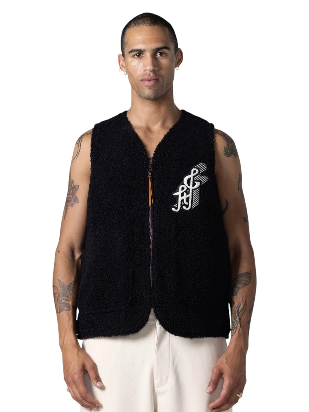 A person wearing the HONOR THE GIFT TEDDY NOTES VEST BLACK, adorned with a white emblem on the left side, white pants, and showcasing multiple tattoos on their arms, stands against a plain white background.