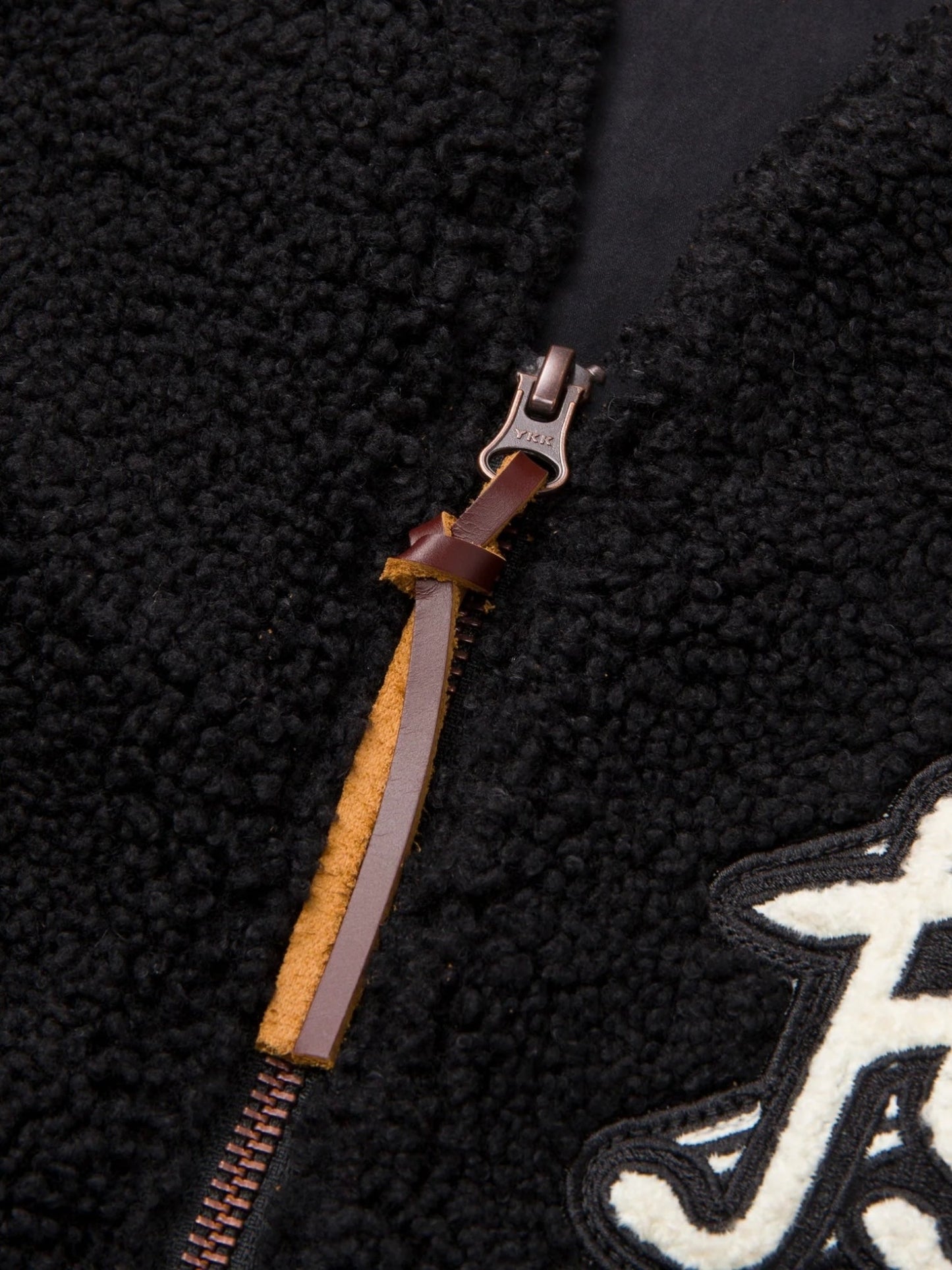 Close-up of the HONOR THE GIFT TEDDY NOTES VEST BLACK, featuring a black fleece material, a brown leather zipper pull, and a white embroidered letter "F" detail.