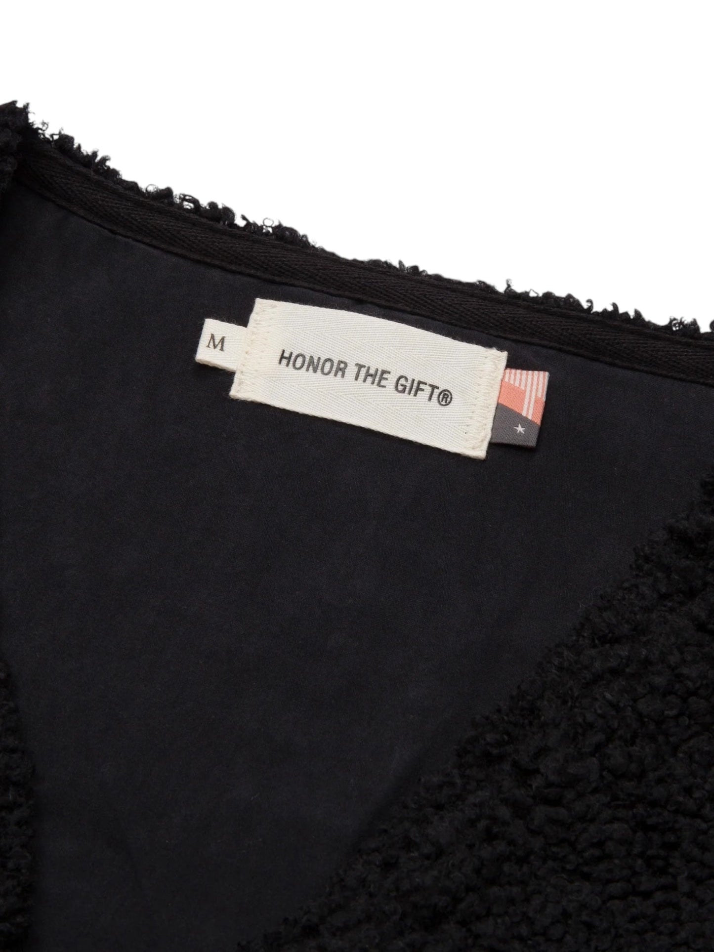 Close-up of a clothing label with the text "Honor the Gift" and an American flag, attached to the neck area of a black textured fabric garment. The label indicates the size as medium (M). The garment depicted is the HONOR THE GIFT TEDDY NOTES VEST in black, from the brand HONOR THE GIFT.