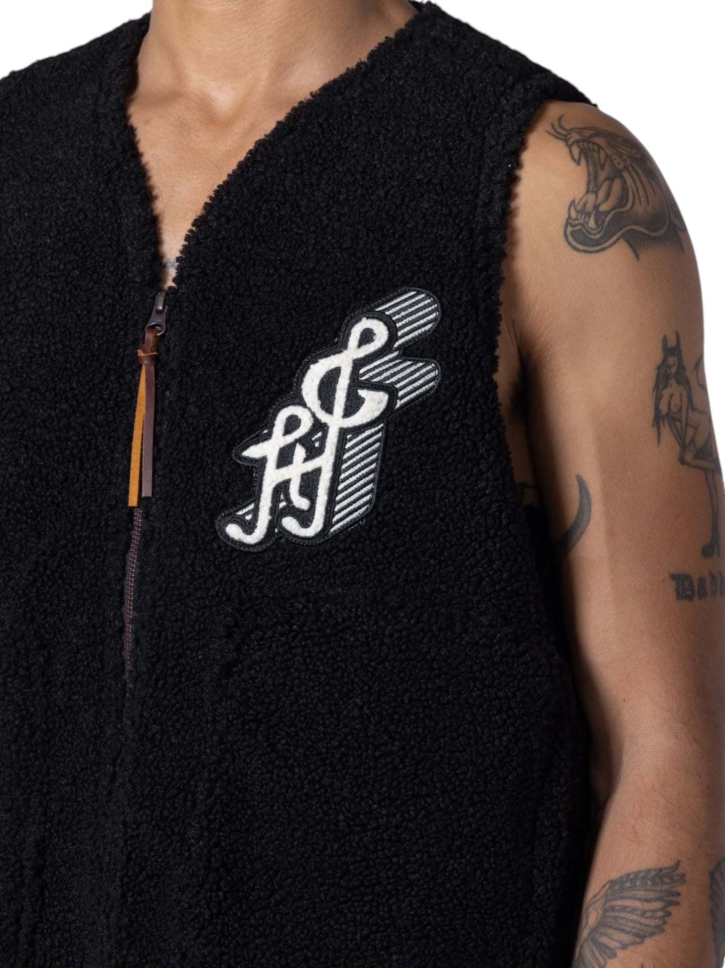 A person wearing an HONOR THE GIFT TEDDY NOTES VEST BLACK, featuring a white embroidered logo on the chest, and showcasing tattoos on their upper right arm.